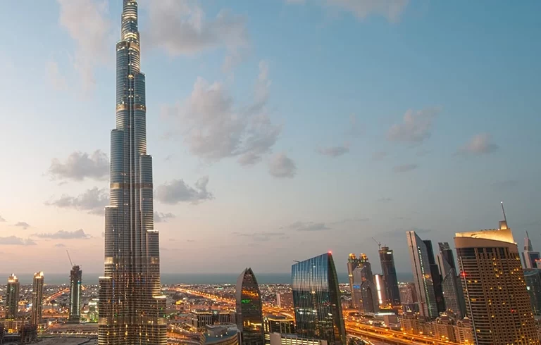 A Dubai cityscape is pictured at sunset. The city is illuminated in the bottom portion of the frame by synthetic light, as natural light lights the top half of the photo. A modern skyscraper points up into the sky in the center of the frame. rare view from the address hotel which is no longer possible.