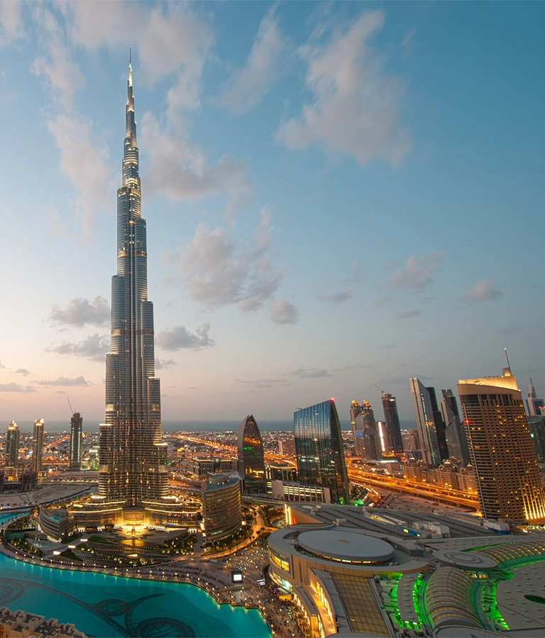 A Dubai cityscape is pictured at sunset. The city is illuminated in the bottom portion of the frame by synthetic light, as natural light lights the top half of the photo. A modern skyscraper points up into the sky in the center of the frame. rare view from the address hotel which is no longer possible.
